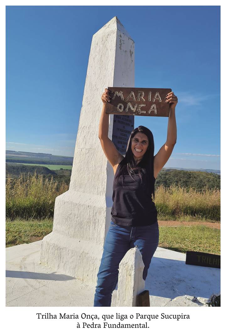 4 z 9:  Uma mulher parda de cabelos pretos,  que traja blusa preta e calça jeans, segura uma placa onde lê-se: Maria Onça talhada na madeira. Ela sorri! Realização: Academia Planaltinense de Letras, Artes e Ciências (APLAC), pelo Ecomuseu Pedra Fundamental e pelo Coletivo Nativo Audiodescrição produzida  pelo Instituto de Promoção das Pessoas com Deficiência Visual Audiodescritora: Elma Lúcia Rodrigues Consultor: Fernando Rodrigues Este projeto é promovido com recursos do Fundo de Apoio a Cultura do DF